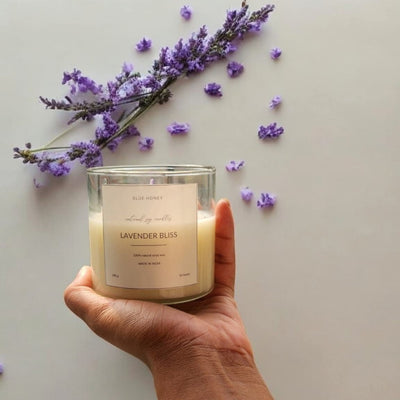 A hand holds a clear glass soy wax candle labeled 'Lavender Bliss' with lavender sprigs and scattered petals.