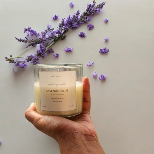 A hand holds a clear glass soy wax candle labeled 'Lavender Bliss' with lavender sprigs and scattered petals.