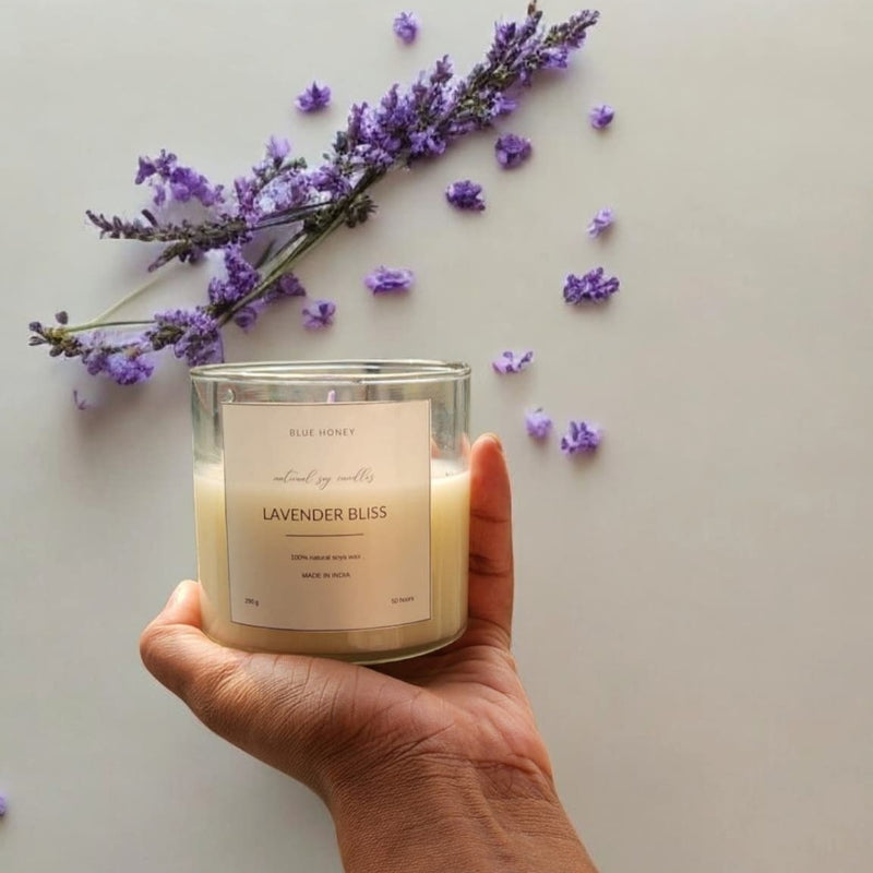 A hand holds a clear glass soy wax candle labeled 'Lavender Bliss' with lavender sprigs and scattered petals.