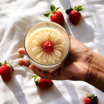 Sun-Kissed Daisy clear glass soy wax candle with a floral design, held by a hand surrounded by strawberries.