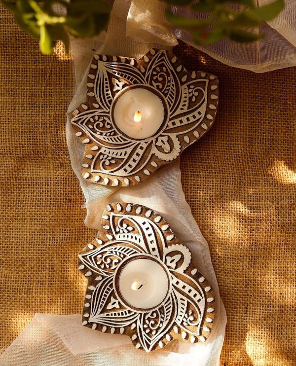 Two handcrafted wooden carved tealight holders with intricate white patterns, lit with candles, on a burlap background.