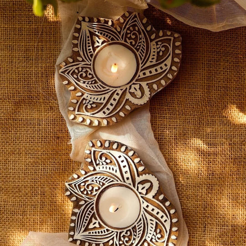 Two handcrafted wooden carved tealight holders with intricate white patterns, lit with candles, on a burlap background.