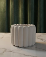 A white scalloped flute Jesmonite scented candle sits on a marble surface in front of green curtains.