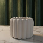 A white scalloped flute Jesmonite scented candle sits on a marble surface in front of green curtains.