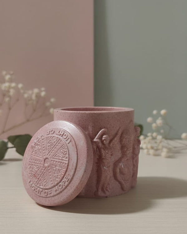 Pink Jesmonite Renaissance Relic Scrolled candle vessel with lid, featuring embossed figures and text.