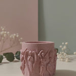 Pink Jesmonite Renaissance Relic candle vessel with scrolled angel details, displayed on a table.