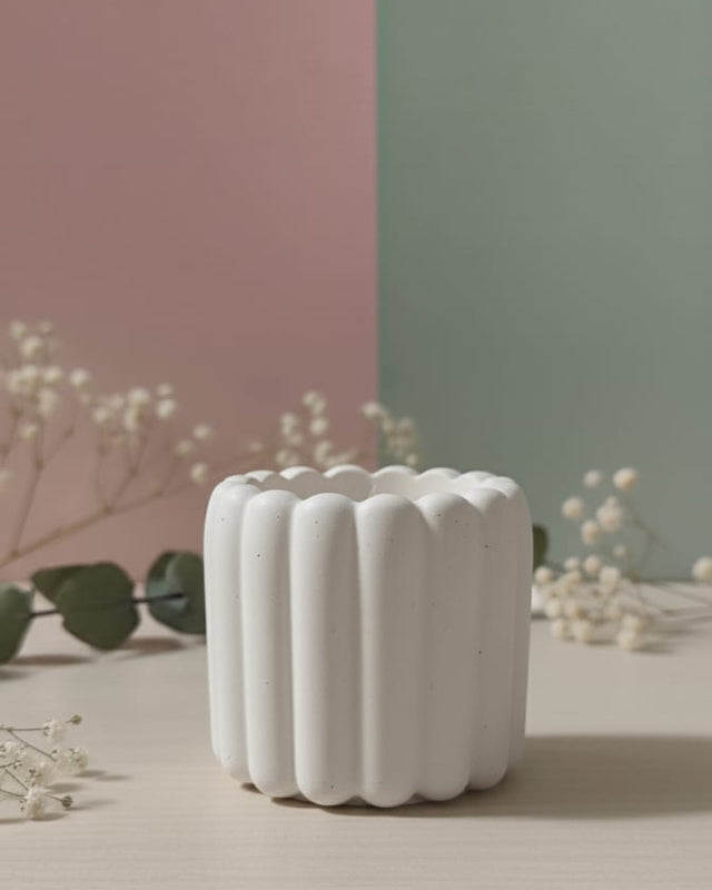 A white scalloped flute Jesmonite scented candle sits on a light-colored surface with delicate white flowers.