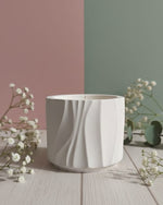 A white Jesmonite Modern Wave Vessel scented candle is displayed from the front on a wooden surface with delicate white flowers.
