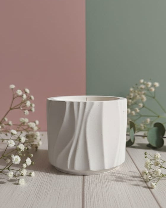 A white Jesmonite Modern Wave Vessel scented candle is displayed from the front on a wooden surface with delicate white flowers.