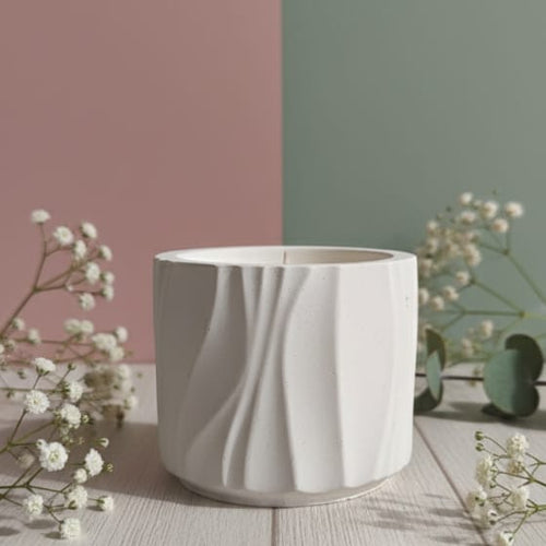A white Jesmonite Modern Wave Vessel scented candle is displayed from the front on a wooden surface with delicate white flowers.