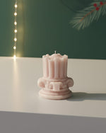 A pink mini Corinthian pillar candle with intricate detailing sits on a white surface, casting a soft shadow.