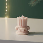 A pink mini Corinthian pillar candle with intricate detailing sits on a white surface, casting a soft shadow.