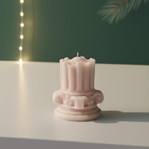 A pink mini Corinthian pillar candle with intricate detailing sits on a white surface, casting a soft shadow.