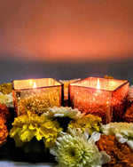 Two scented cuboid-shaped glass jar candles in gold and rose gold, surrounded by flowers.
