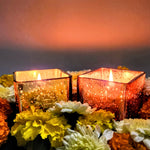 Two scented cuboid-shaped glass jar candles in gold and rose gold, surrounded by flowers.