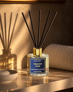 A reed diffuser bottle with a blue and white label reading 'BLUE HONEY WELCOME HOME SIGNATURE COLLECTION REED DIFFUSER' sits on a warm-toned wooden surface with diffuser reeds extending upward. The scene is styled in a bathroom-like setting with a towel, bowl, and softly lit background.
