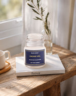 A clear glass jar candle labeled 'Blue Honey Orchid Veil Signature Collection' sits on a white book on a rustic wooden table, with a white mug at left and a glass vase with dried stems in the background near a window.