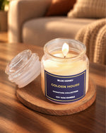A soy wax candle in a clear glass jar sits on a round wooden coaster on a wooden table, with its lid placed beside it. The background shows a softly blurred living room setting with a sofa and cushions.