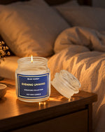 A lit soy wax candle in a clear glass jar sits on a wooden bedside table next to its matching glass lid, with a softly blurred bed in the background. The label reads 'BLUE HONEY EVENING UNWIND SIGNATURE COLLECTION SOY WAX CANDLE'.
