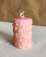 A 4-inch tall soy wax pillar candle in baby pink with gold-dusted floral embellishments.