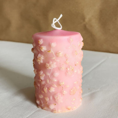 A 4-inch tall soy wax pillar candle in baby pink with gold-dusted floral embellishments.