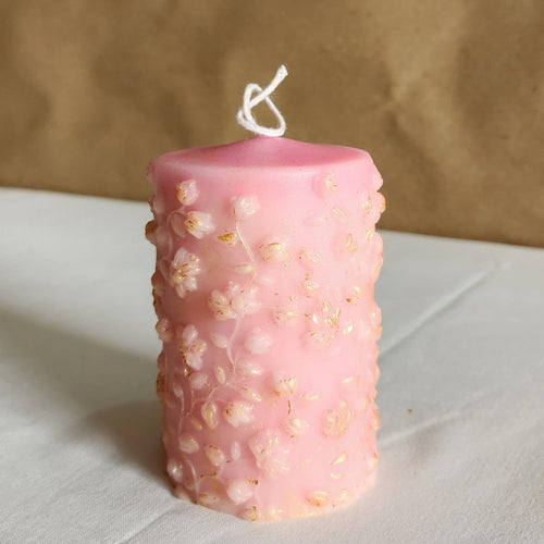 A 4-inch tall soy wax pillar candle in baby pink with gold-dusted floral embellishments.