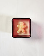 A festive gingerbread man candle with gold accents sits in a red glass jar, perfect for Christmas gifting.