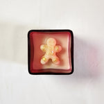 A festive gingerbread man candle with gold accents sits in a red glass jar, perfect for Christmas gifting.