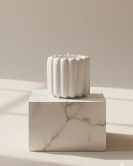 A white scalloped flute scented Jesmonite candle sits on a marble pedestal, casting soft shadows.