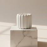 A white scalloped flute scented Jesmonite candle sits on a marble pedestal, casting soft shadows.