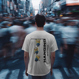 Back view of a white unisex t-shirt featuring three Minions hanging from a bar with the text 'Hanging By A Thread'.