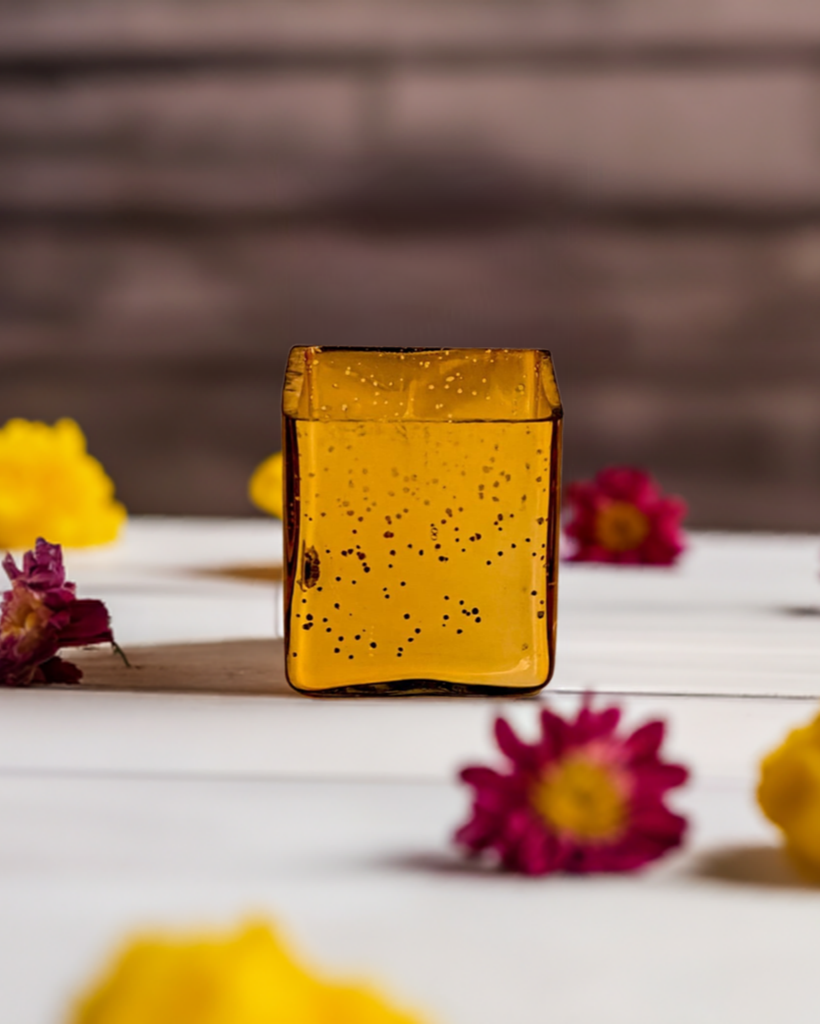 A cuboid-shaped gold glass jar candle with a glittery finish, surrounded by flowers.