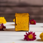 A cuboid-shaped gold glass jar candle with a glittery finish, surrounded by flowers.