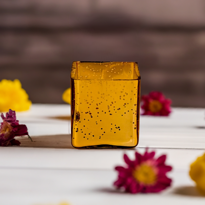 A cuboid-shaped gold glass jar candle with a glittery finish, surrounded by flowers.
