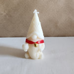 A white gnome-shaped soy wax pillar candle with a red ribbon and gold bell, perfect for Christmas gifts.