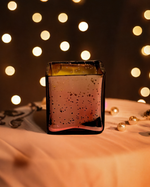 A rose gold cuboid-shaped glass jar candle with a speckled finish sits on a draped cloth, with bokeh lights in the background.