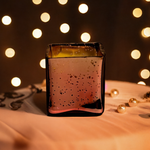 A rose gold cuboid-shaped glass jar candle with a speckled finish sits on a draped cloth, with bokeh lights in the background.