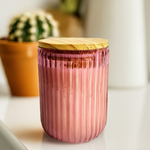Ribbed Radiance scented glass jar candle in pink, featuring a ribbed design and wooden lid, displayed on a white surface.