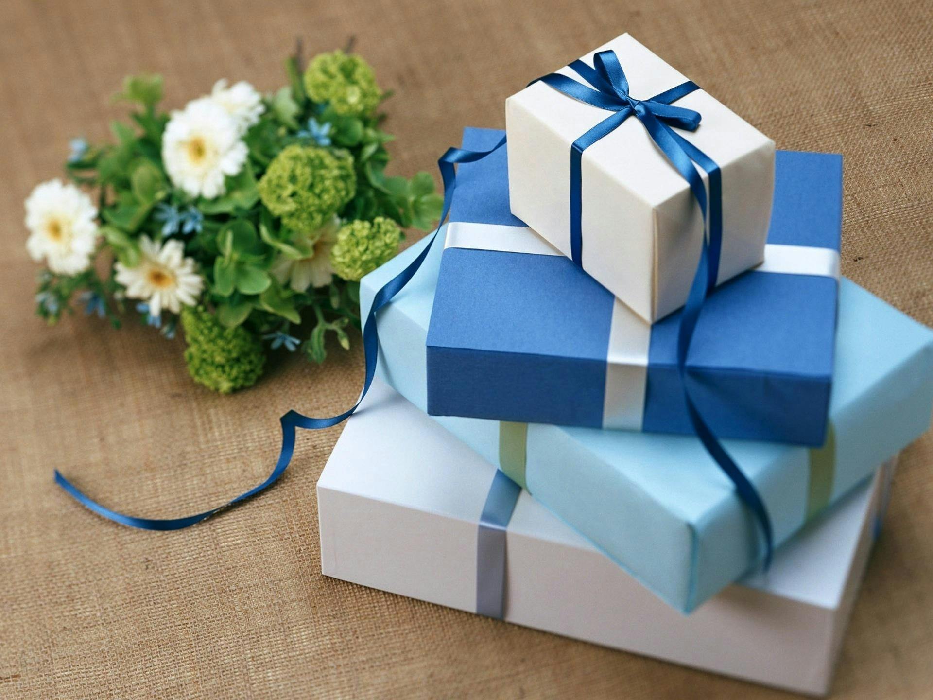 Stack of blue and white gift boxes tied with blue ribbon, next to a floral arrangement on a burlap surface.