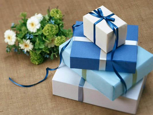 Stack of blue and white gift boxes tied with blue ribbon, next to a floral arrangement on a burlap surface.