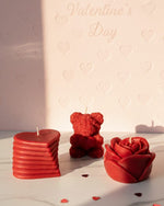 Three red sculpted Valentine's Day candles: a heart, a teddy bear, and a rose, on a marble surface.