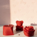 Three red sculpted Valentine's Day candles: a heart, a teddy bear, and a rose, on a marble surface.