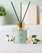 A clear glass reed oil diffuser with a gold cap and a light green label, surrounded by jasmine flowers.