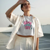 A woman wears a white 'Girls Who Lift' crop top and white pants, shielding her eyes from the sun.