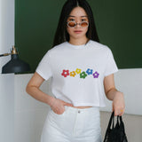 A young woman models the white Rainbow Bloom Crop Top, featuring a vibrant rainbow flower graphic, perfect for casual summer style.