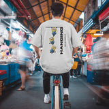 Back view of a white unisex Minions 'Hanging By A Thread' t-shirt worn by a person cycling through a market.