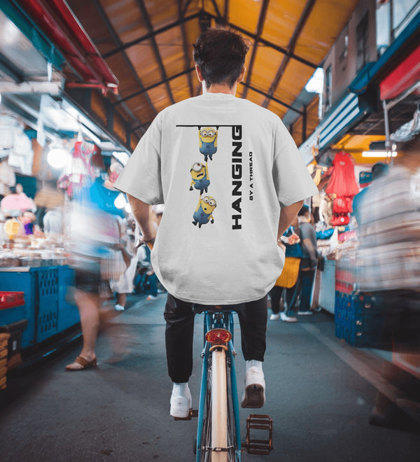 Back view of a white unisex Minions 'Hanging By A Thread' t-shirt worn by a person cycling through a market.