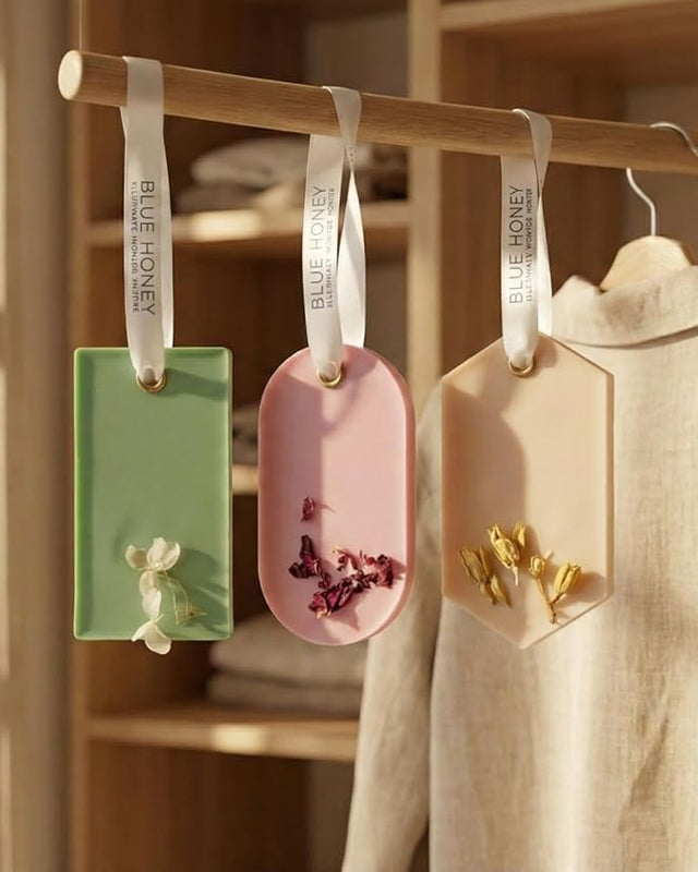 Three handcrafted natural beeswax wax sachets in green, pink, and beige with dried flowers, hanging on a wooden rod.